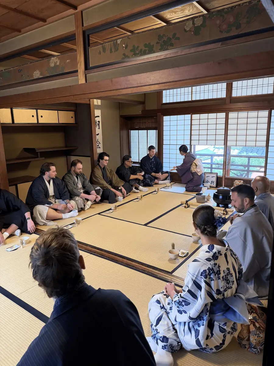 Guests seated on tatami for a tea ceremony, a host in a blue and white kimono