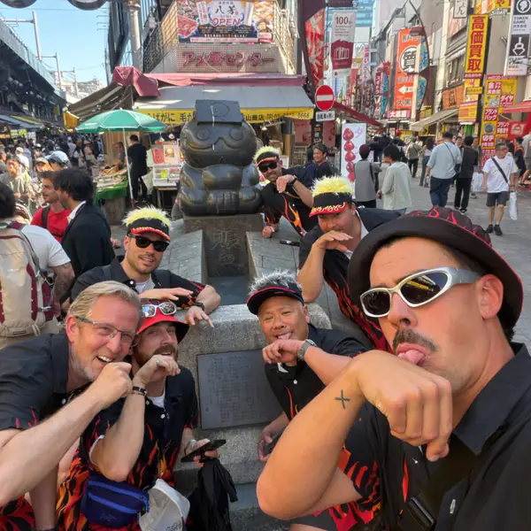 A race team in sunglasses and headbands, posing by a statue on a busy Ueno street